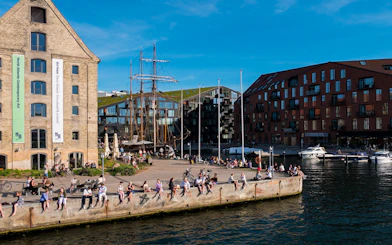 People relaxing by the waterfront in Copenhagen with historic buildings and boats in view.