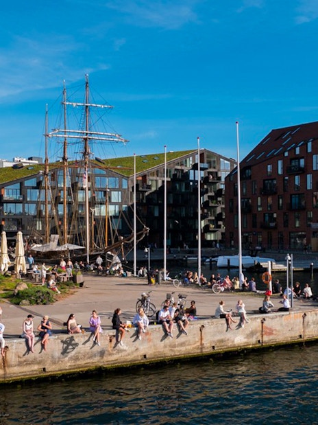 People relaxing by the waterfront in Copenhagen with historic buildings and boats in view.