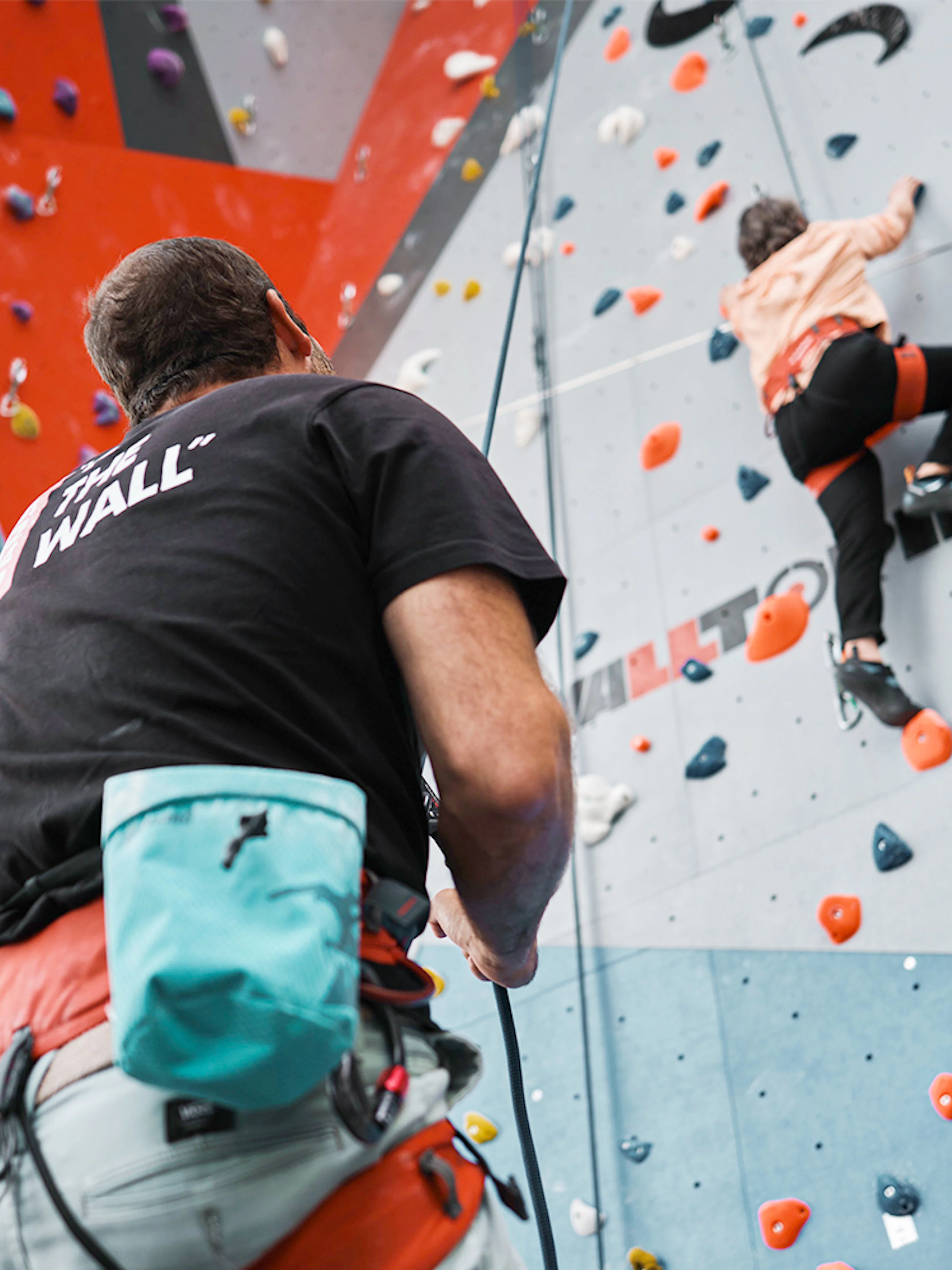 Person climbing indoor rock wall with colorful holds, guided by instructor.