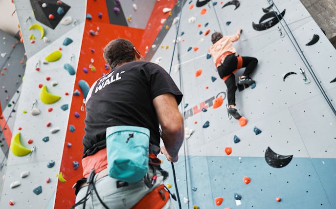 Person climbing indoor rock wall with colorful holds, guided by instructor.