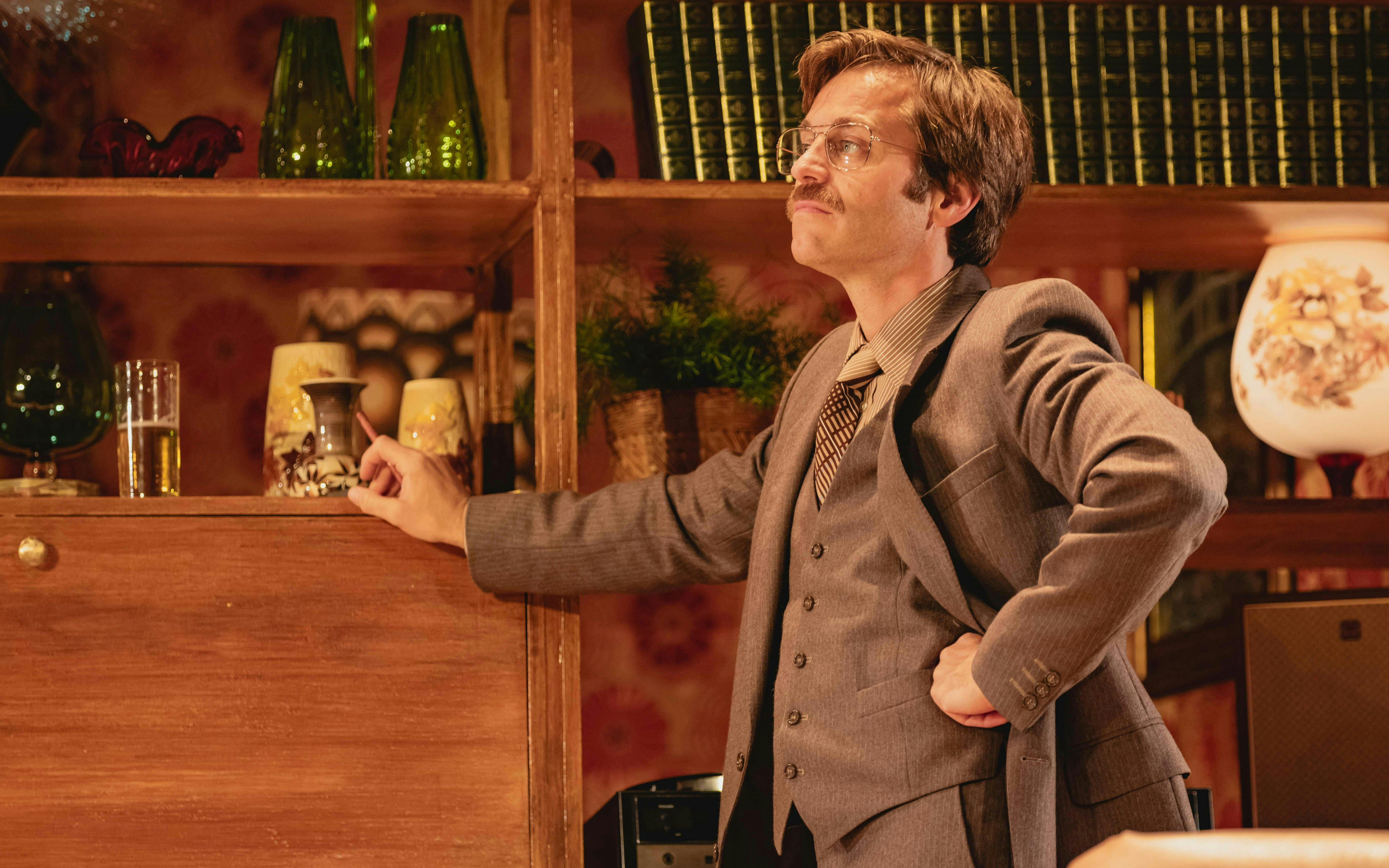 Stage scene from Abigail's Party, Westend, featuring a man in a suit with a vintage decor backdrop.