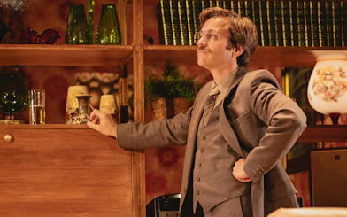 Stage scene from Abigail's Party, Westend, featuring a man in a suit with a vintage decor backdrop.