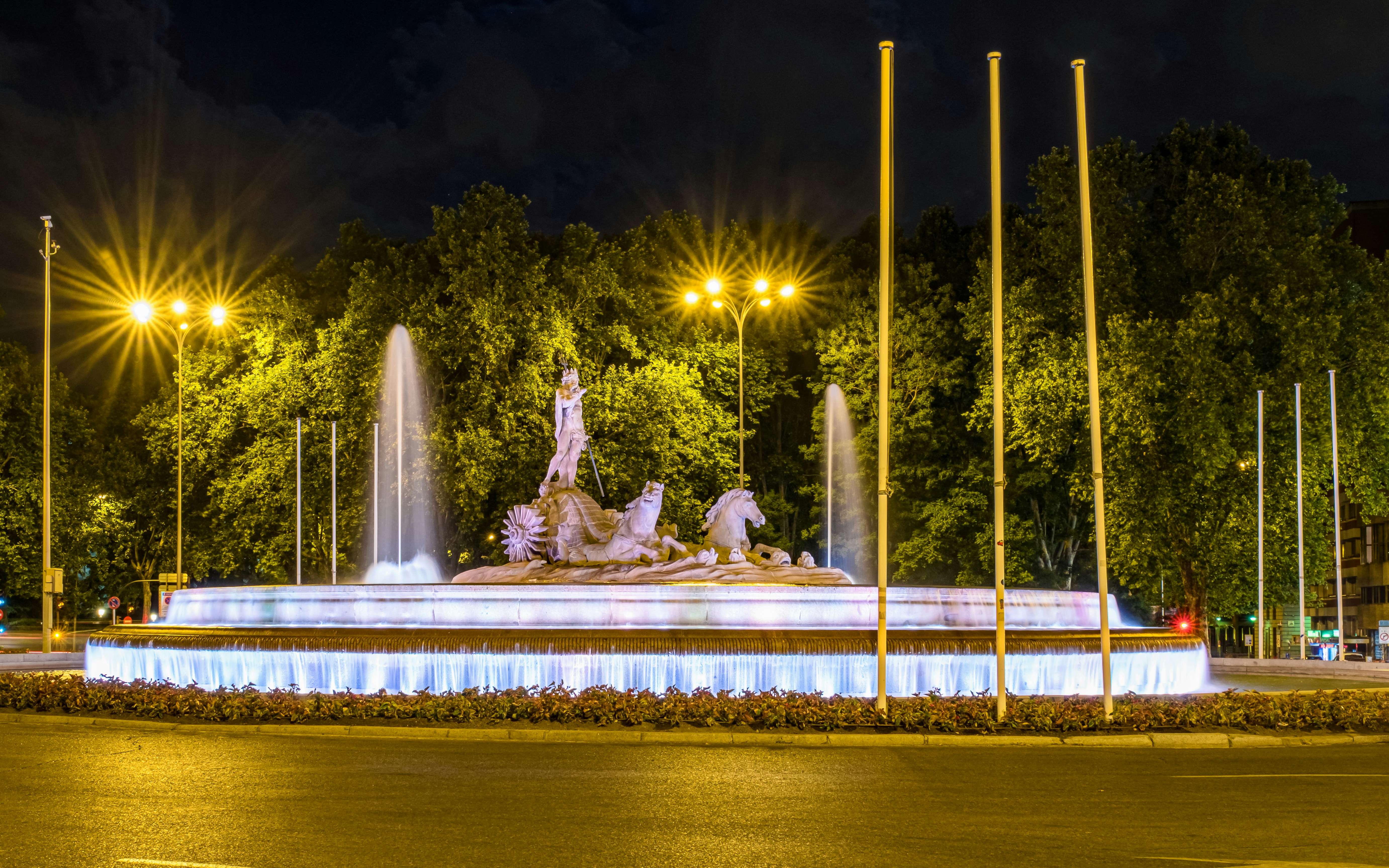Neptune Fountain illuminated at night in Madrid, surrounded by trees and streetlights.