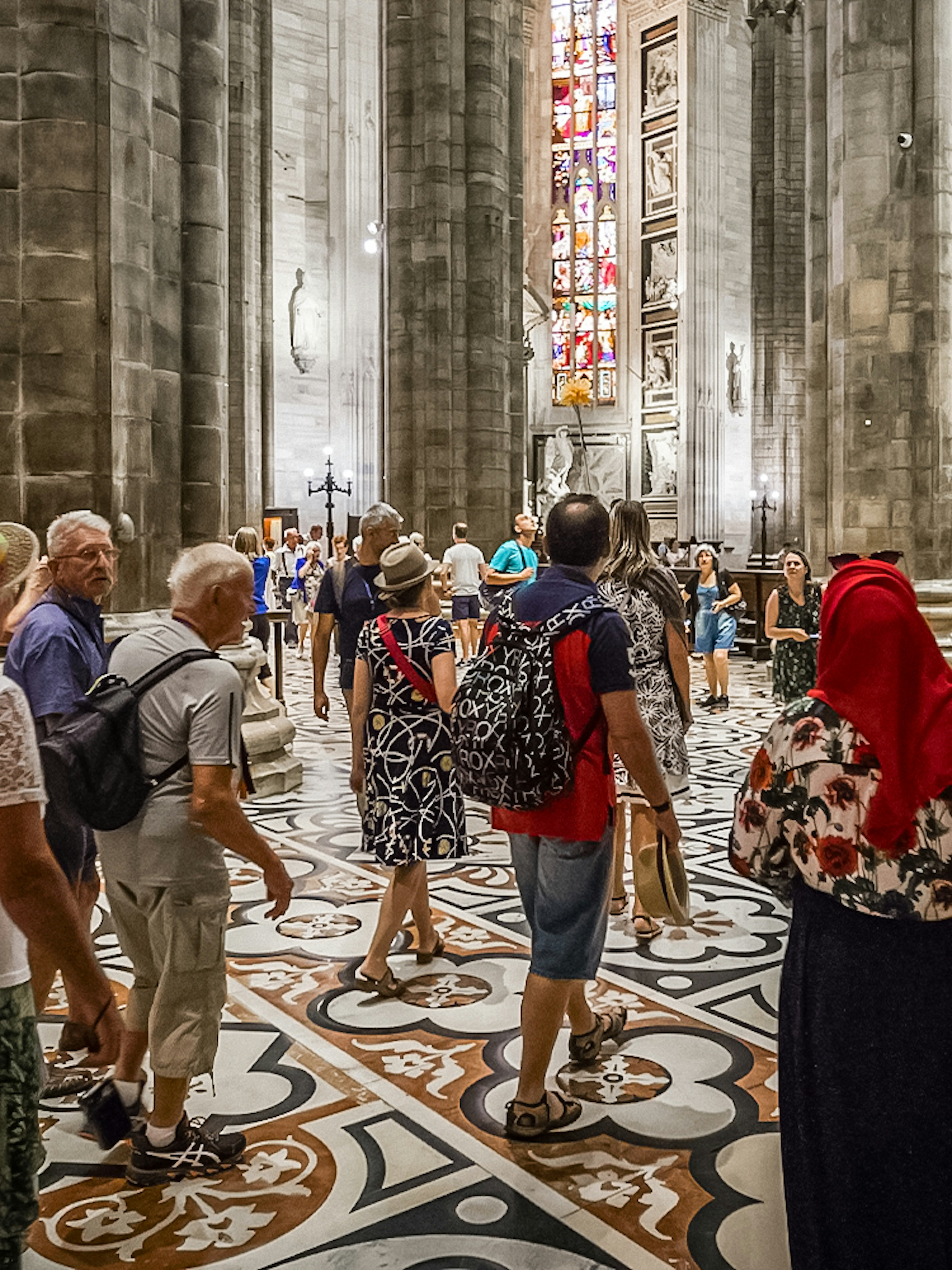 Tourists inside Duomo Cathedral, Milan, admiring Gothic architecture and stained glass.