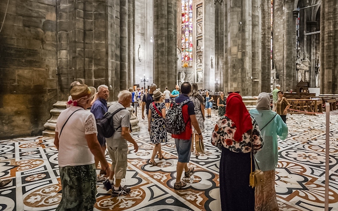 Tourists inside Duomo Cathedral, Milan, admiring Gothic architecture and stained glass.