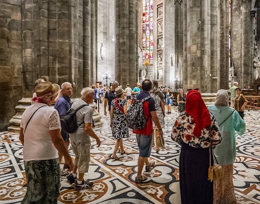 Tourists inside Duomo Cathedral, Milan, admiring Gothic architecture and detailed interior design.