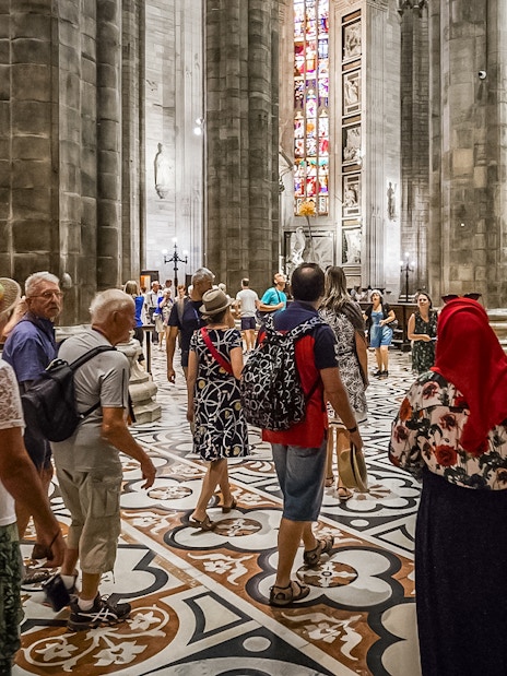 Tourists inside Duomo Cathedral, Milan, admiring Gothic architecture and stained glass.
