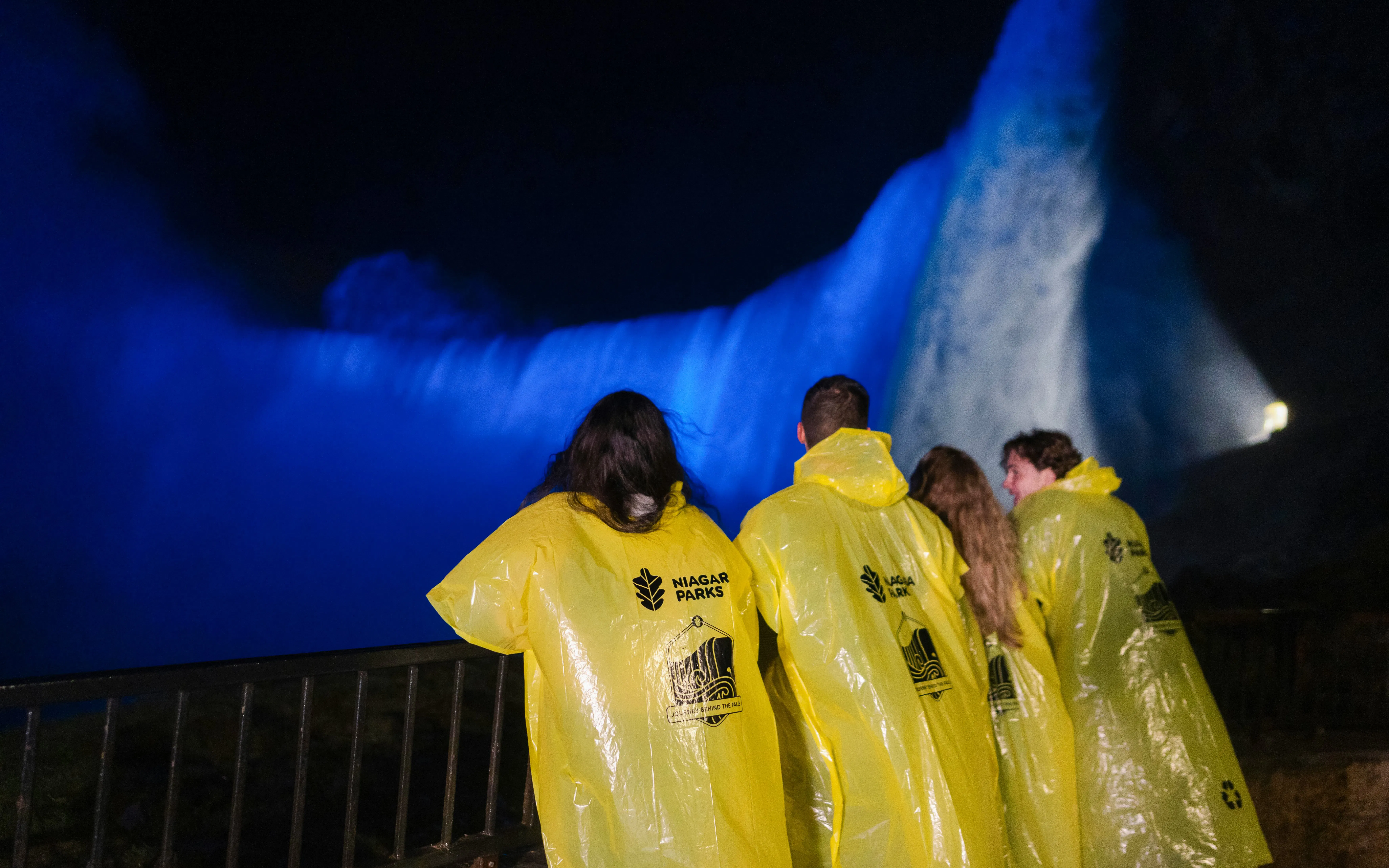 Visitors in yellow ponchos view illuminated Niagara Falls at night from a railing.