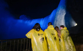Visitors in yellow ponchos view illuminated Niagara Falls at night from a railing.