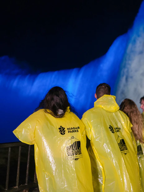 Visitors in yellow ponchos view illuminated Niagara Falls at night from a railing.