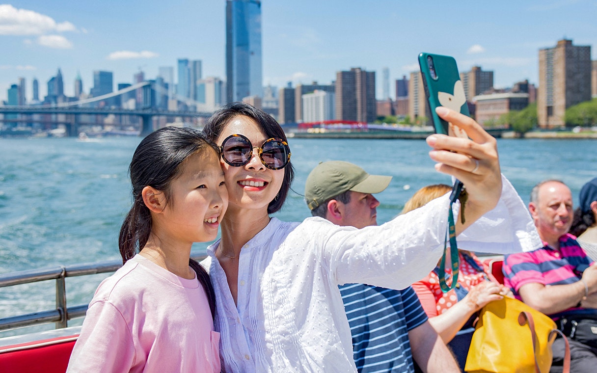 Tourists taking a selfie on a boat with New York City skyline and Brooklyn Bridge in the background.