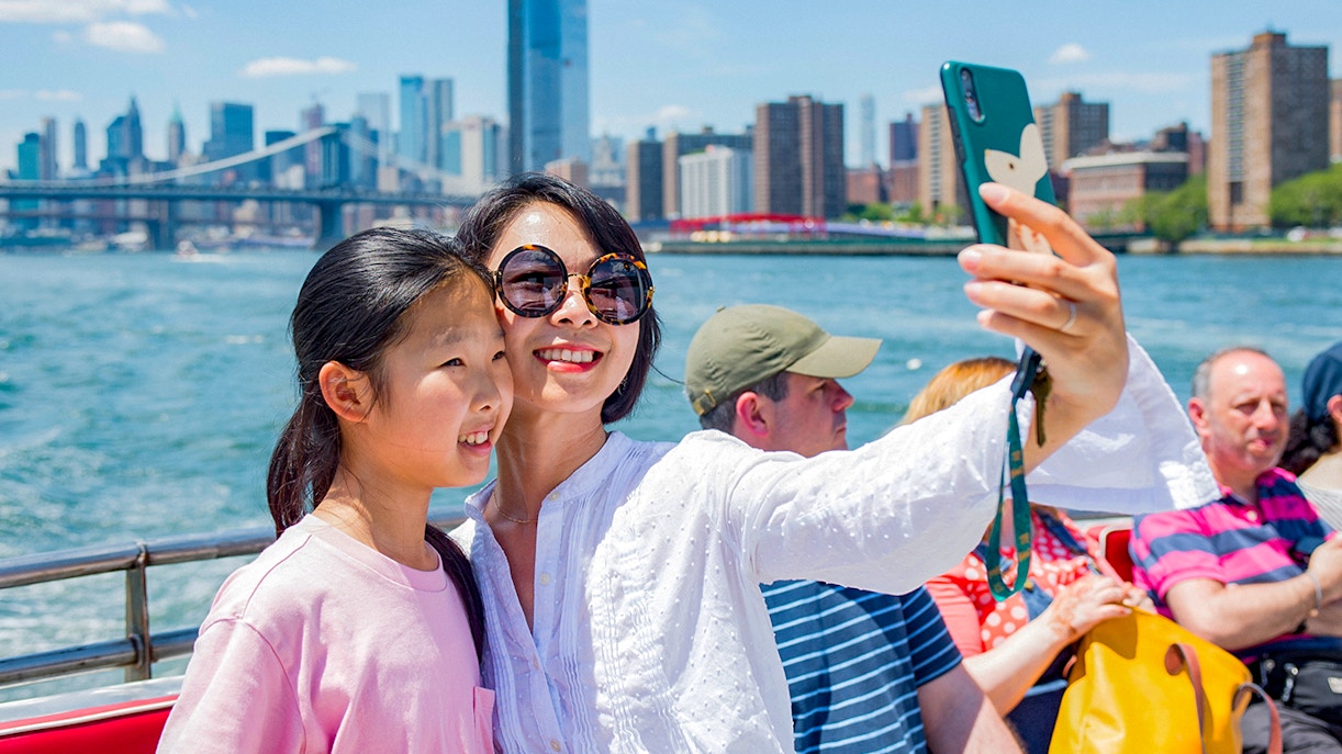 Tourists taking a selfie on a boat with New York City skyline and Brooklyn Bridge in the background.
