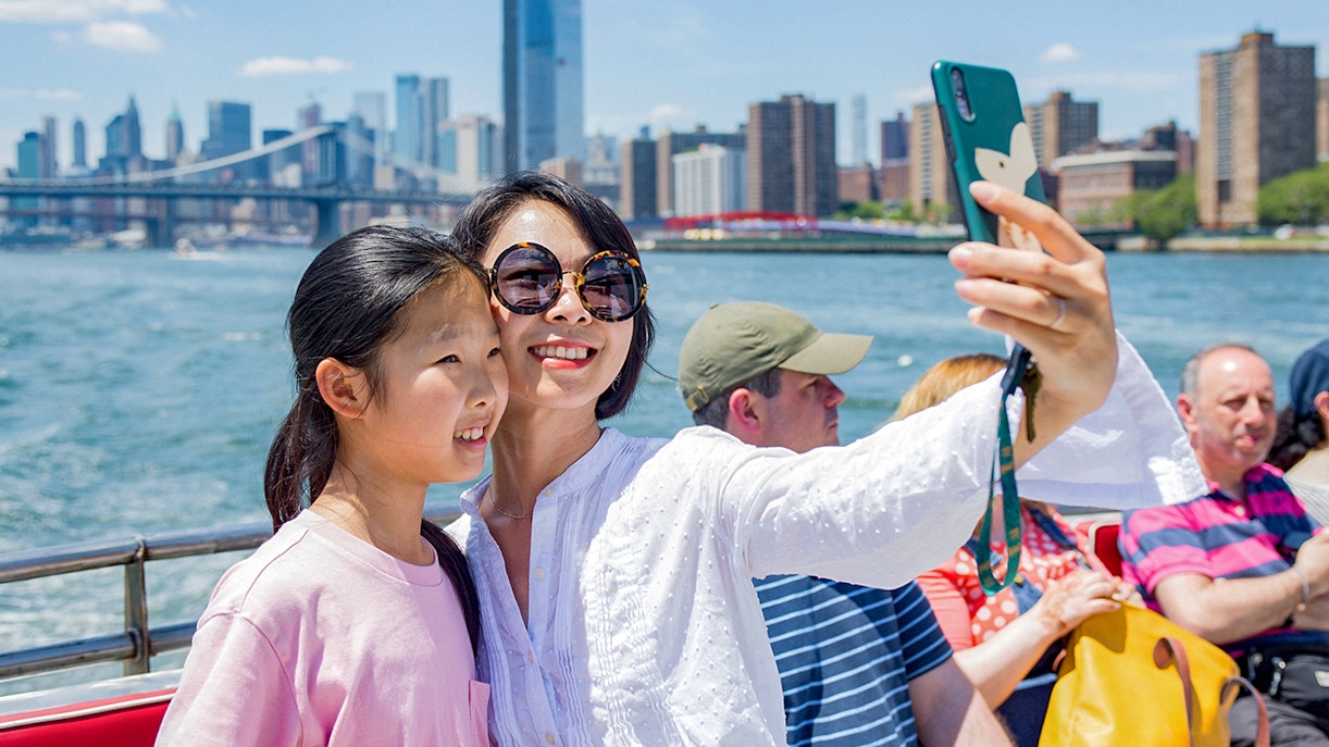 Tourists taking a selfie on a boat with New York City skyline and Brooklyn Bridge in the background.