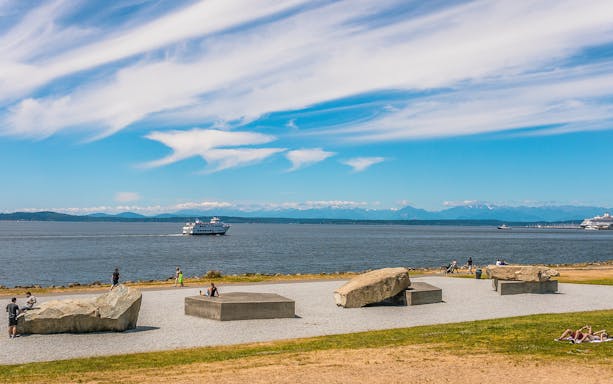 Seattle waterfront view with ferry and people relaxing near sculptures.