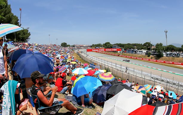 Crowd with umbrellas watching Formula 1 race from general admission at Barcelona circuit.