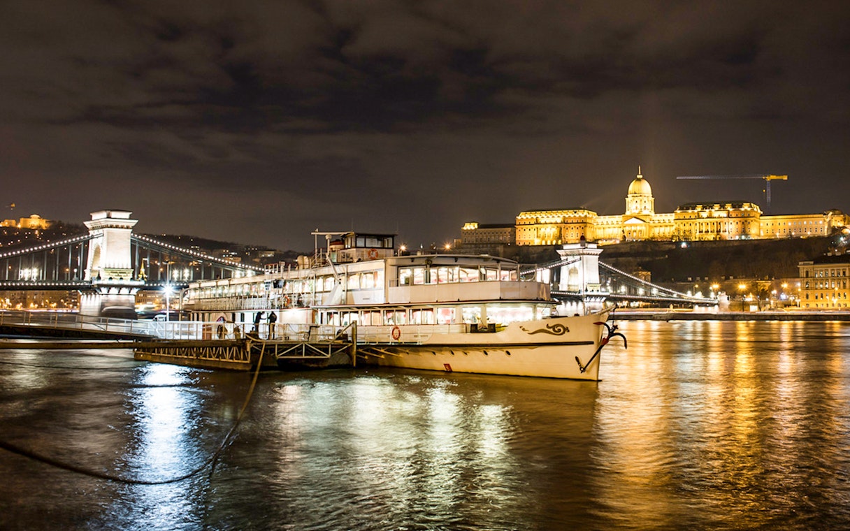 Sightseeing cruise on the Danube River with Budapest's Chain Bridge and Buda Castle illuminated at night.