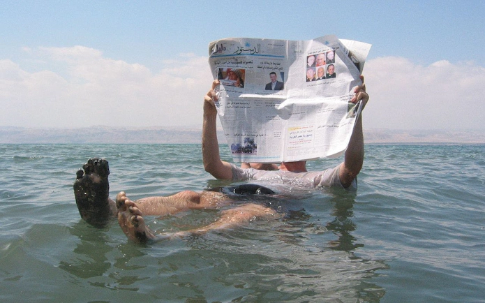 Person floating in the Dead Sea reading a newspaper, with distant hills in the background.