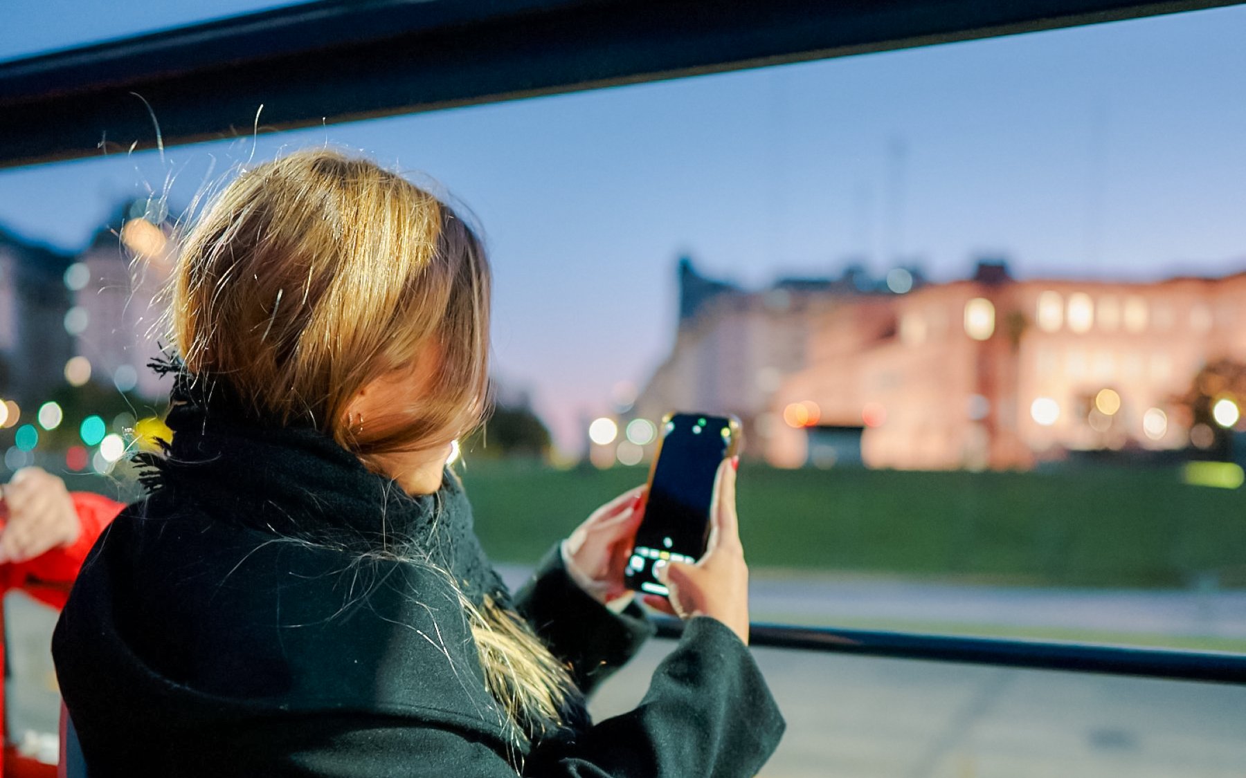 Tourist on Buenos Aires night bus tour capturing cityscape with smartphone.