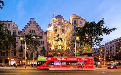 Red tour bus passing Casa Batlló in Barcelona at dusk.