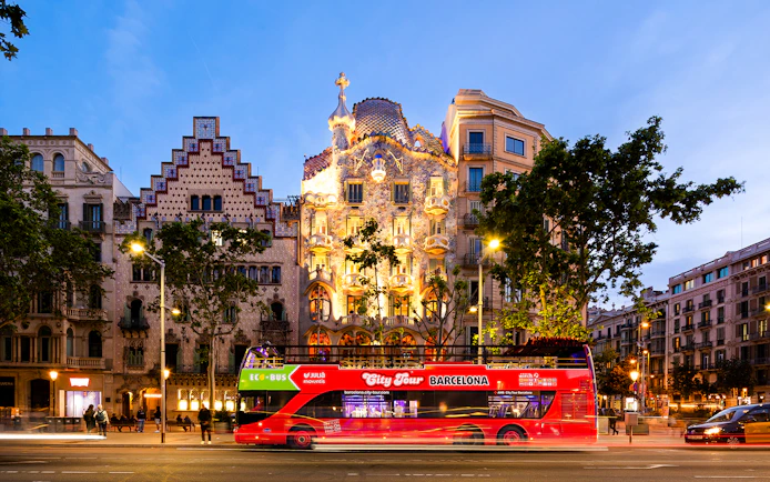 Red tour bus passing Casa Batlló in Barcelona at dusk.