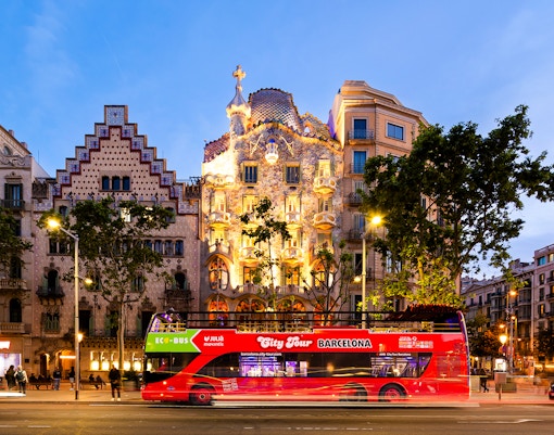 Red tour bus passing Casa Batlló in Barcelona at dusk.