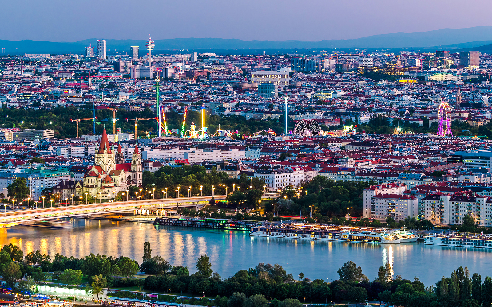 Aerial view of Vienna with skyscrapers, historic buildings, and riverside promenade at dusk.