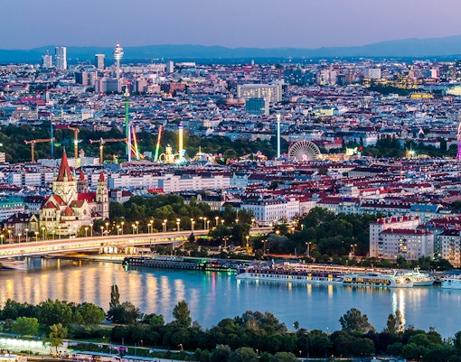 Aerial view of Vienna with skyscrapers, historic buildings, and riverside promenade at dusk.