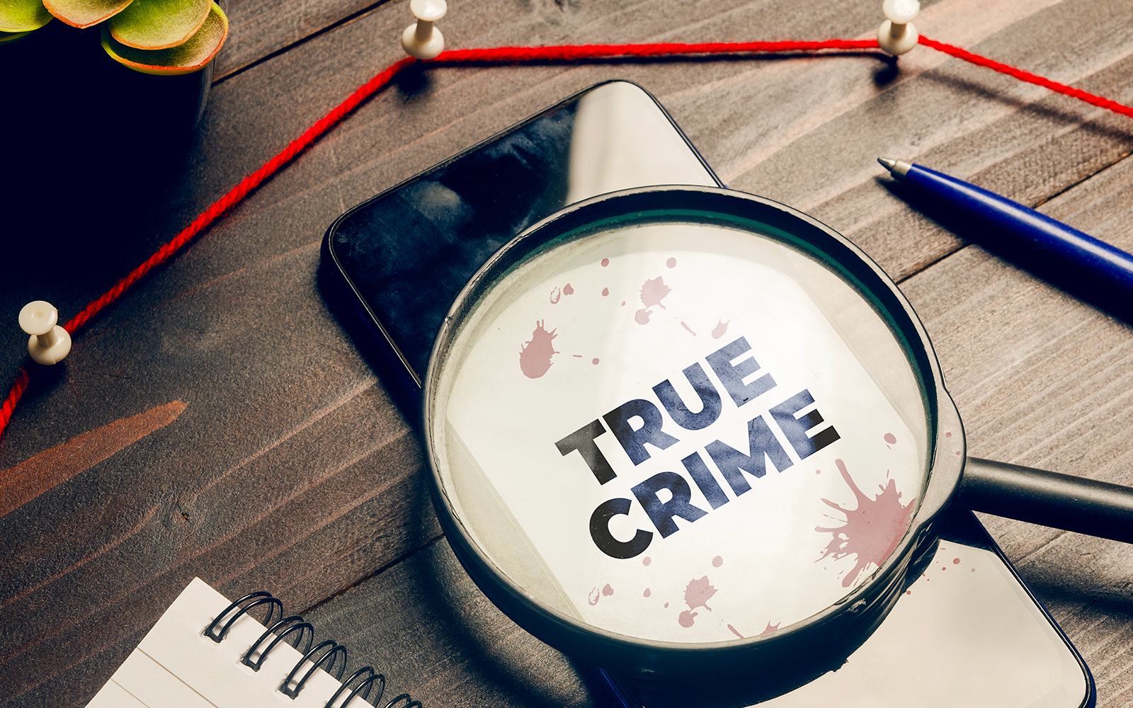 Magnifying glass over "True Crime" text with red string and pushpins on a desk.
