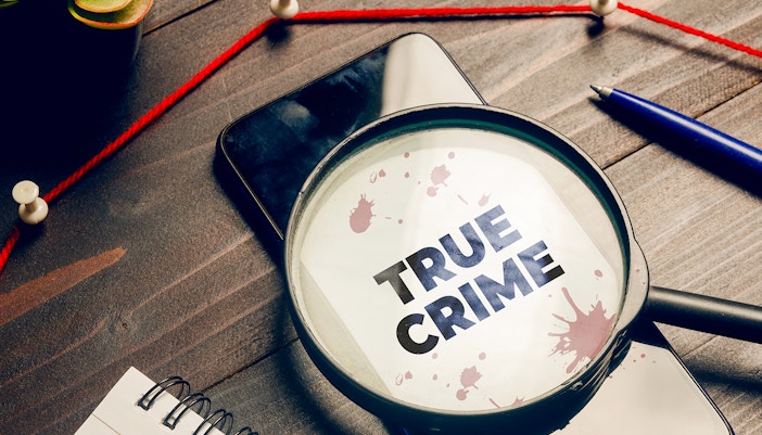 Magnifying glass over "True Crime" text with red string and pushpins on a desk.