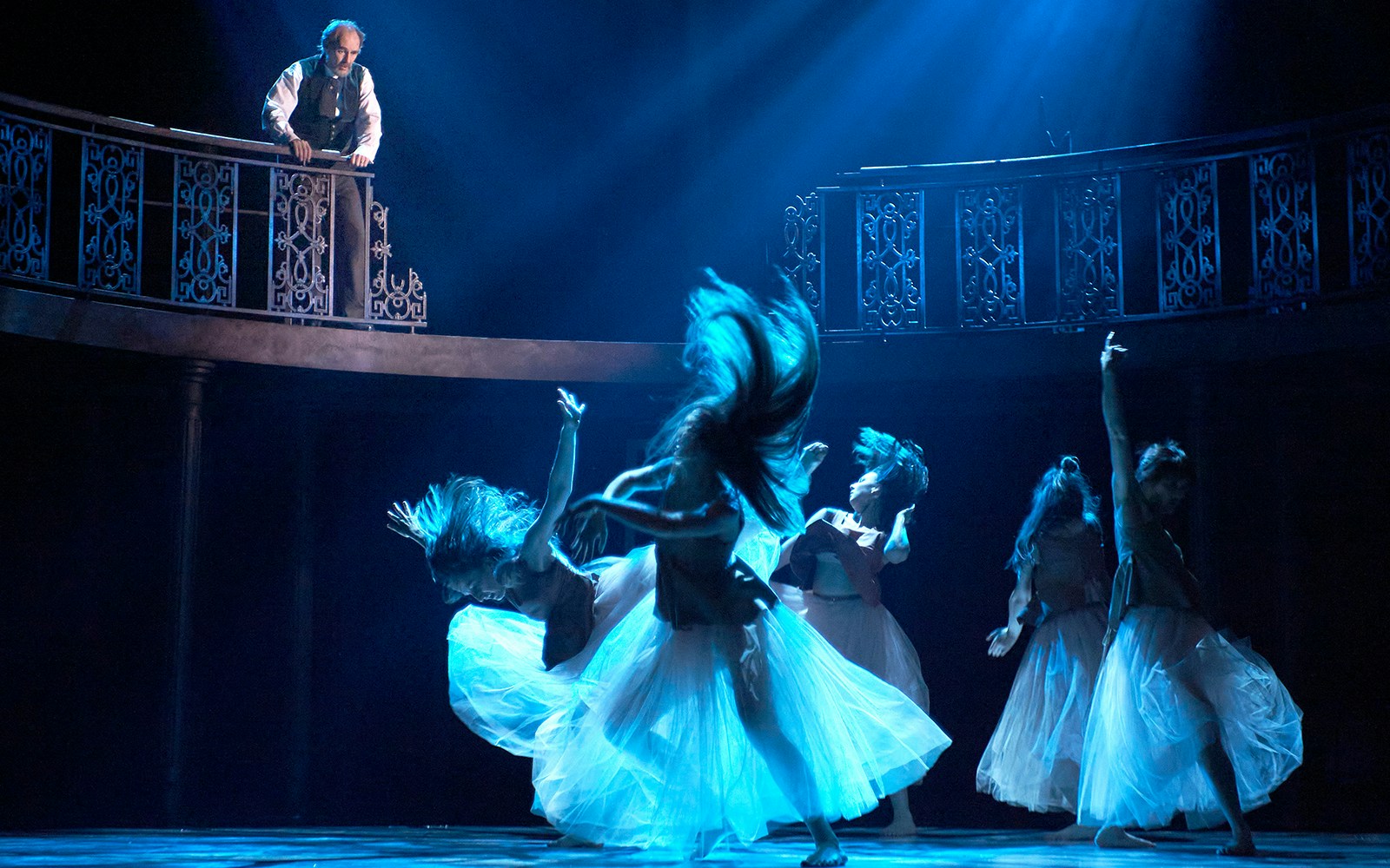Dancers perform under blue light as a man observes from a balcony in a theatrical scene.