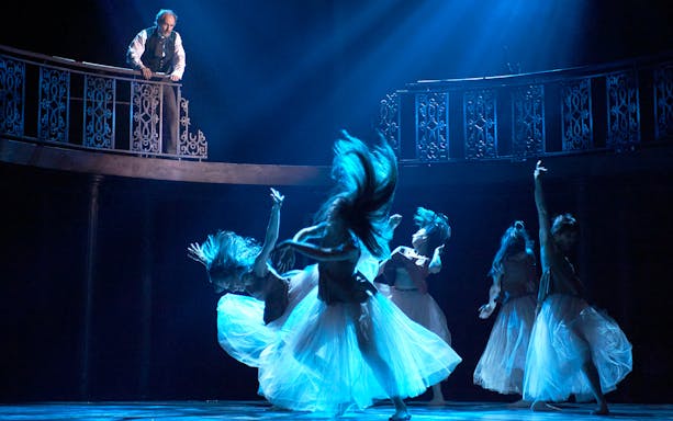 Dancers perform under blue light as a man observes from a balcony in a theatrical scene.