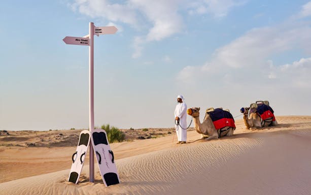 Camel safari guide with camels in Dubai desert near signpost and sandboards.