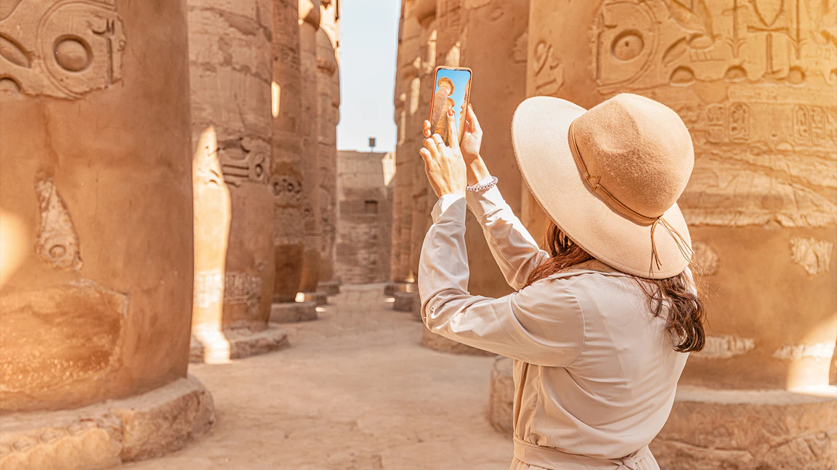 Visitor using phone for audio tour at Karnak Temple, Egypt, surrounded by ancient columns.