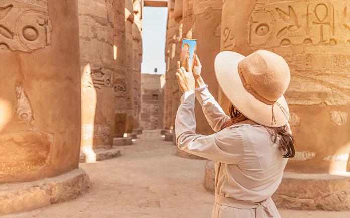 Visitor using phone for audio tour at Karnak Temple, Egypt, surrounded by ancient columns.