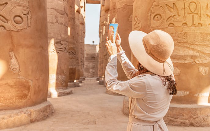 Visitor using phone for audio tour at Karnak Temple, Egypt, surrounded by ancient columns.