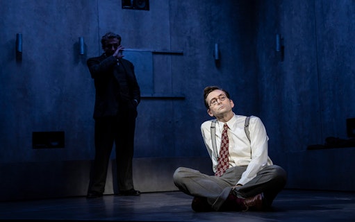 Theater performance scene with a man sitting on the floor, another standing in shadow.