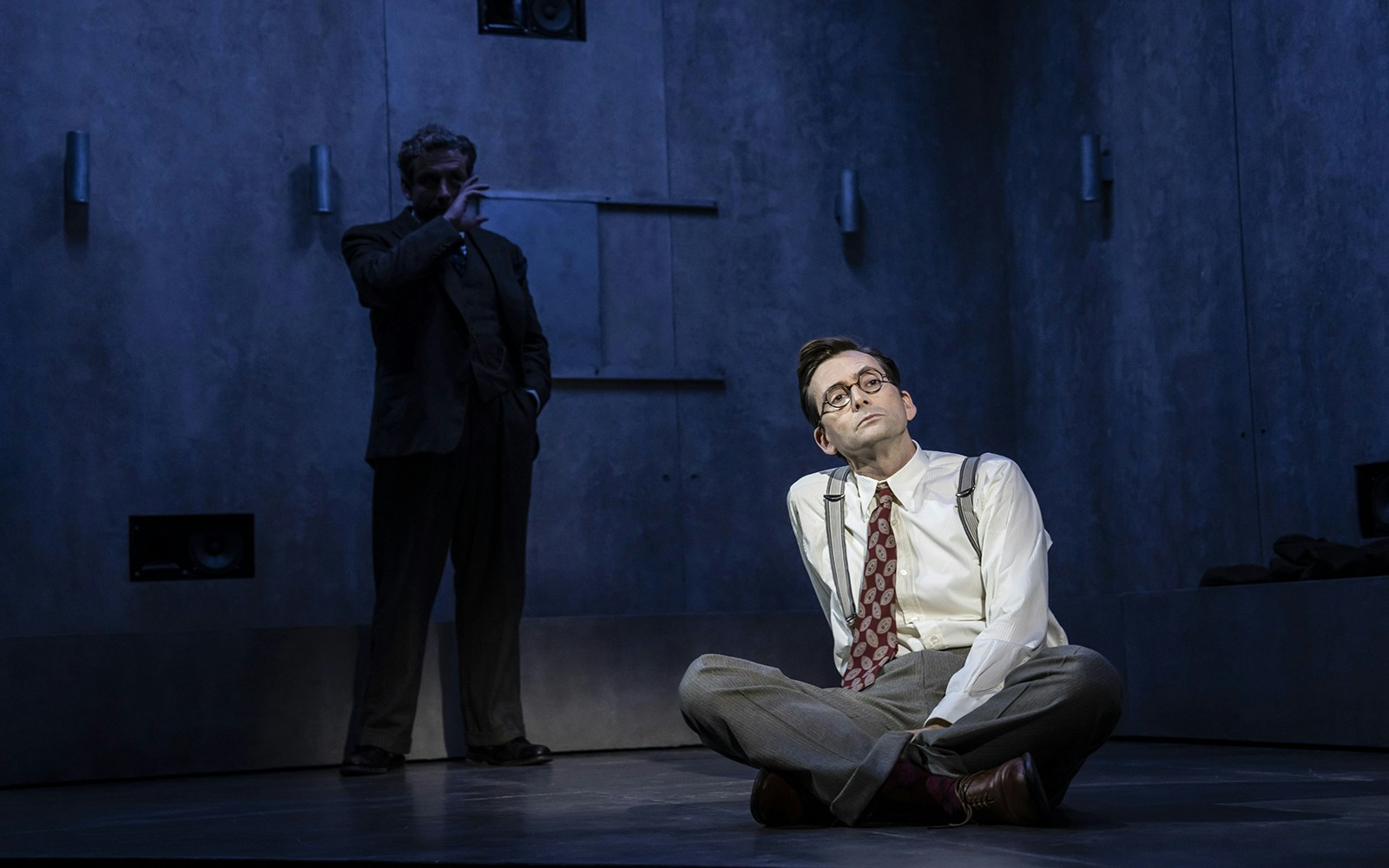 Theater performance scene with a man sitting on the floor, another standing in shadow.
