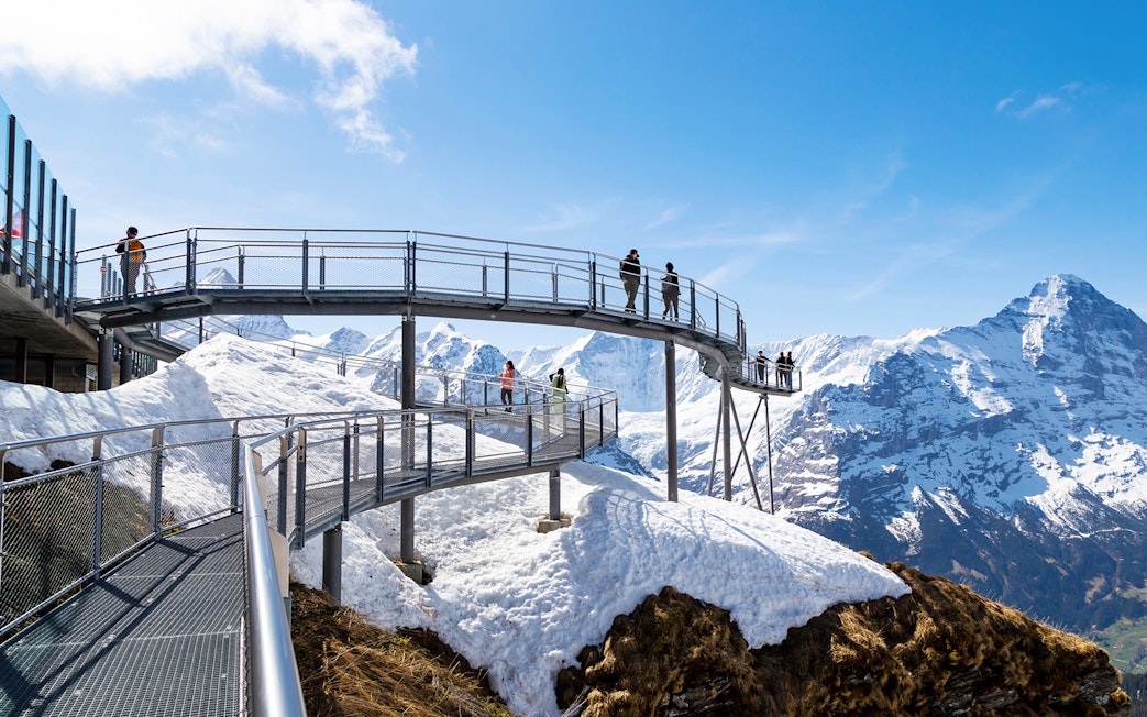 Visitors on a metal walkway with snowy mountain views in Grindelwald, Switzerland.