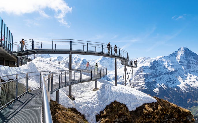 Visitors on a metal walkway with snowy mountain views in Grindelwald, Switzerland.