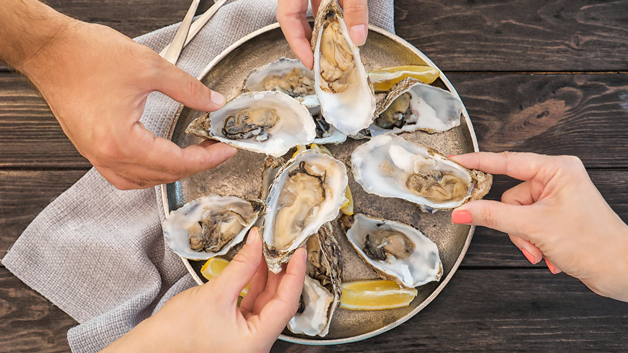 Hands holding fresh oysters on a plate with lemon wedges on a wooden table.