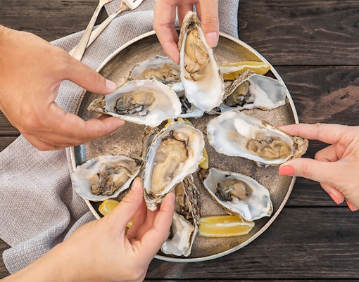 Hands holding fresh oysters on a plate with lemon wedges on a wooden table.