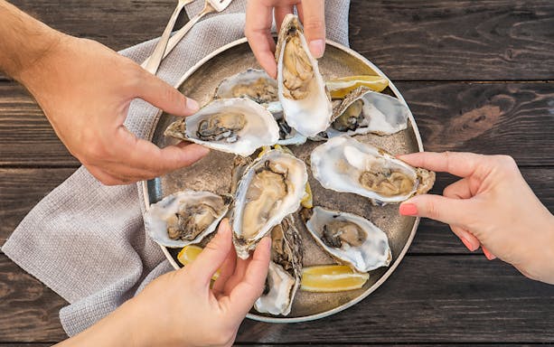 Hands holding fresh oysters on a plate with lemon wedges on a wooden table.