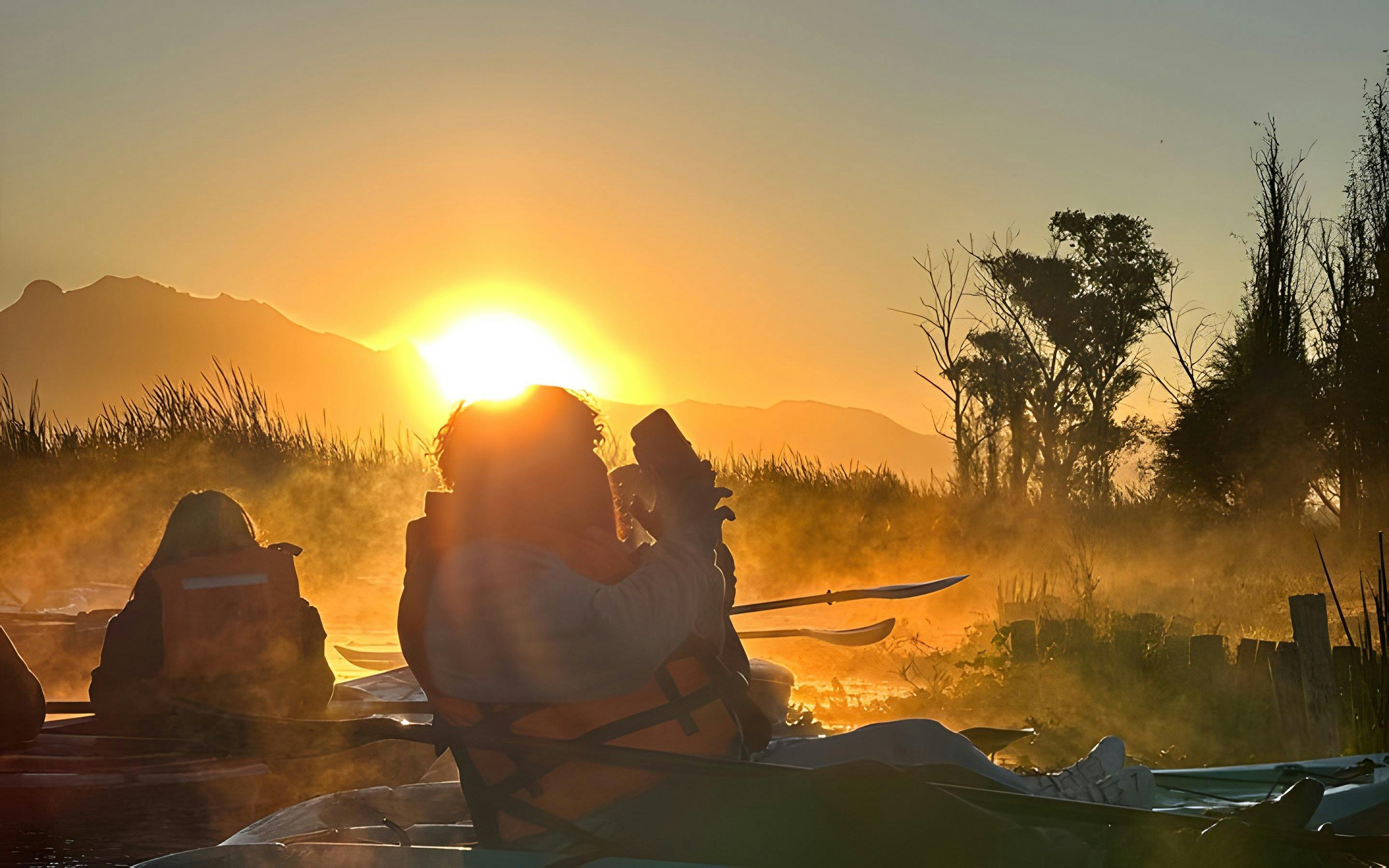 Kayakers in Xochimilco canals at sunrise with mist and silhouetted trees.