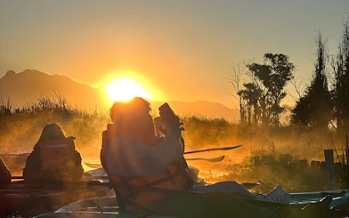 Kayakers in Xochimilco canals at sunrise with mist and silhouetted trees.