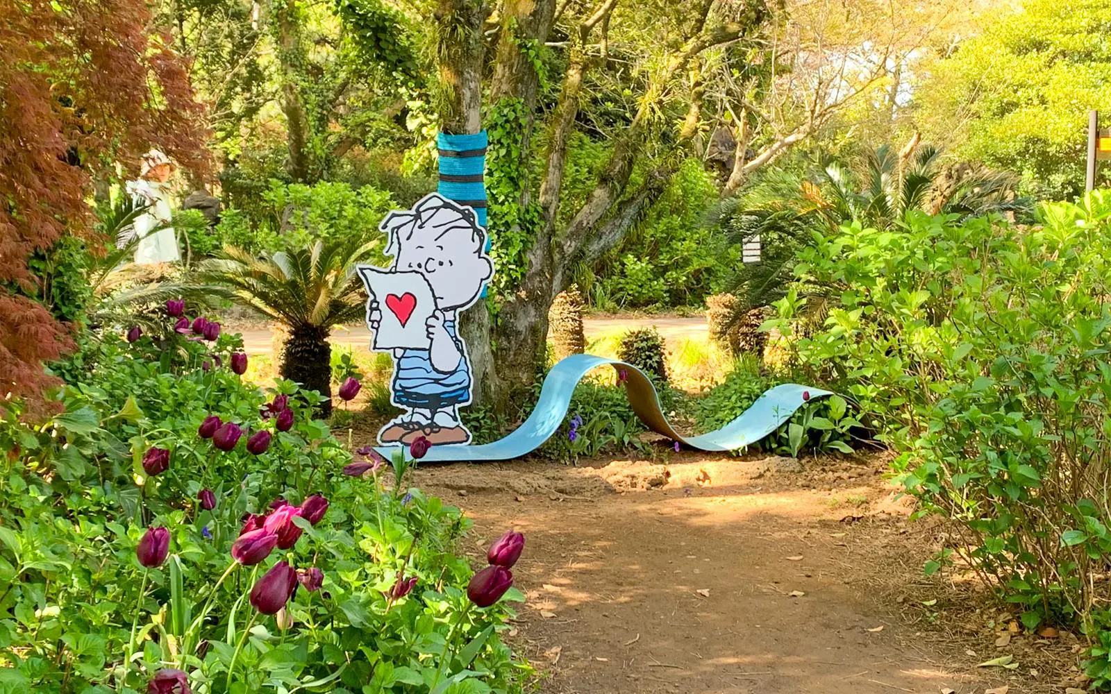 Character holding heart card in Snoopy Garden, Jeju, surrounded by tulips and trees.