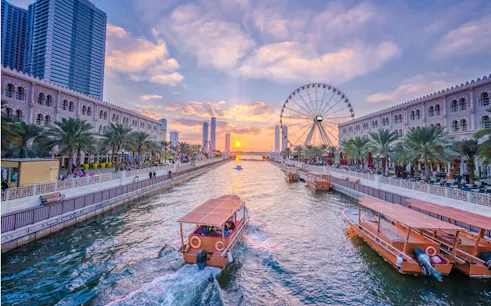 Waterfront view of Sharjah with boats, palm-lined promenade, and Ferris wheel at sunset.