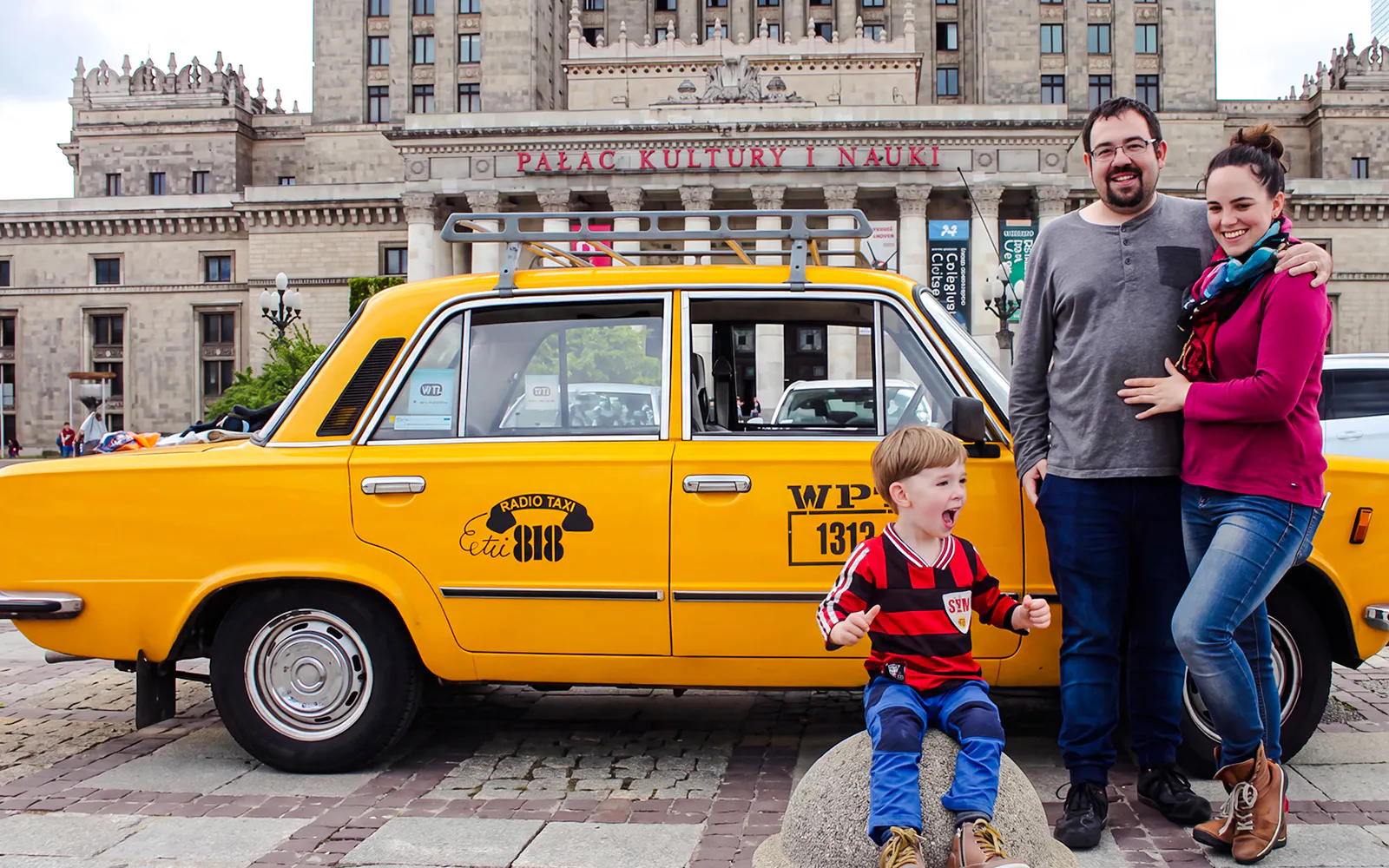 Family posing with yellow Fiat 125p taxi in front of Palace of Culture and Science, Warsaw.