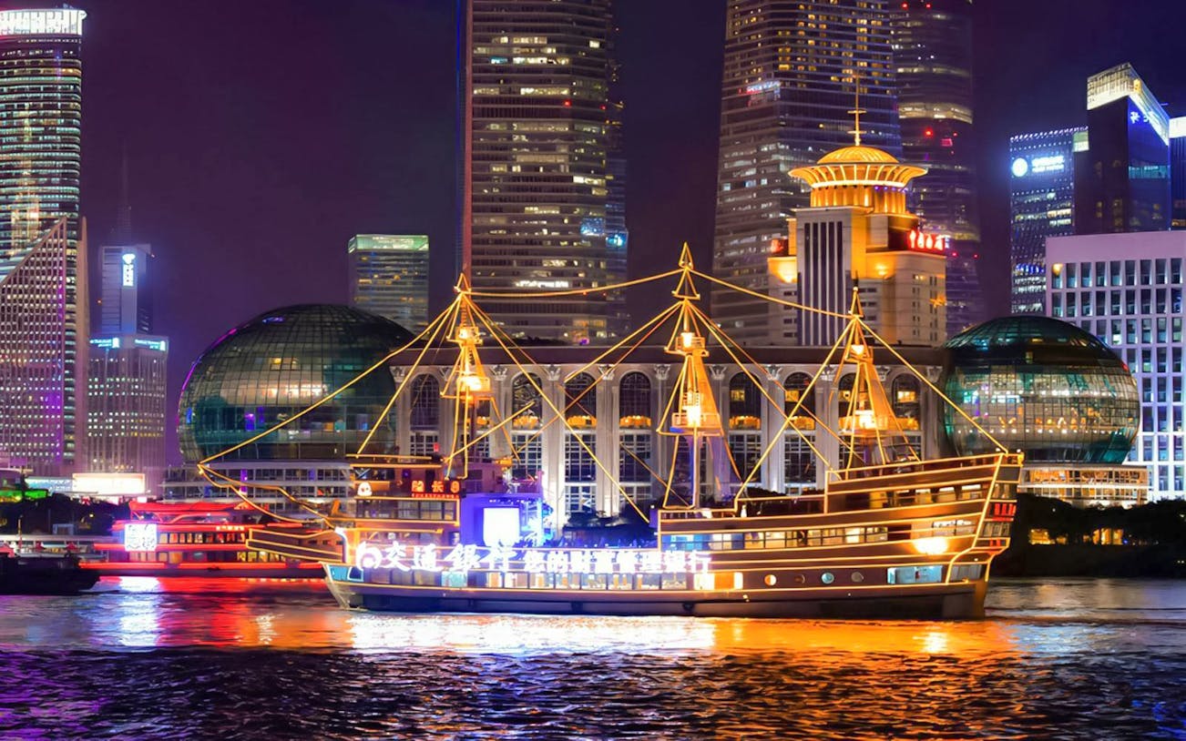 Traditional-style cruise ship illuminated on the Huangpu River, Shanghai skyline in background.