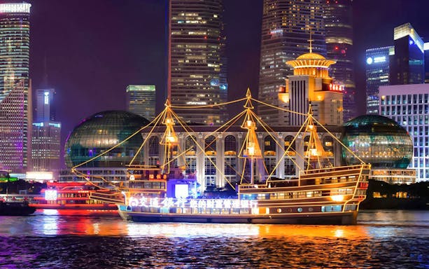 Traditional-style cruise ship illuminated on the Huangpu River, Shanghai skyline in background.