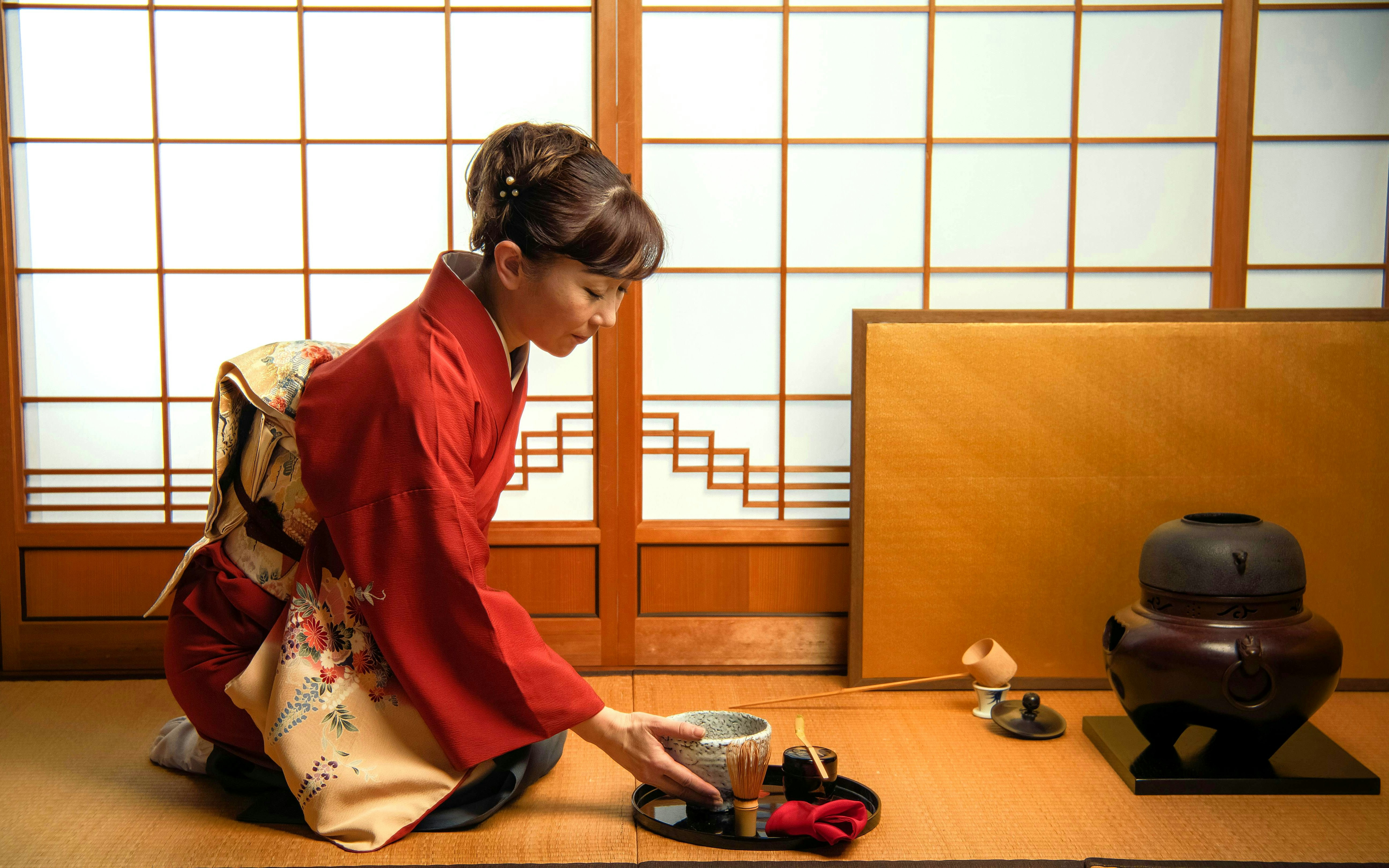 Asian woman in red kimono preparing tea ceremony with traditional utensils.